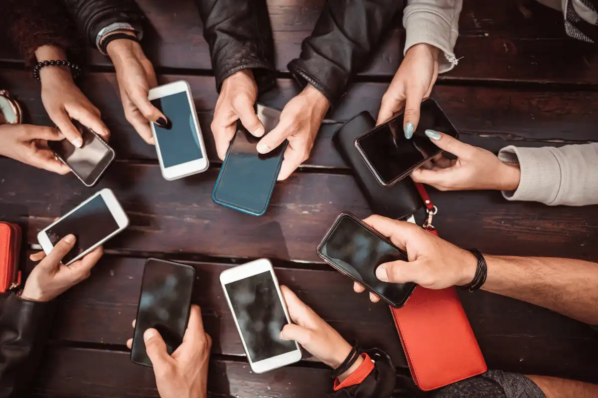 Várias mãos segurando smartphones sobre uma mesa de madeira. A imagem retrata a interação social moderna com a tecnologia móvel.