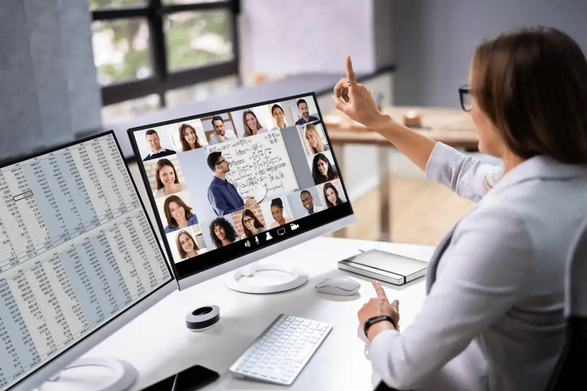 Pessoa participando de videoconferência em dois monitores, um apresentando uma aula e outro com vários participantes, ambiente de trabalho moderno.