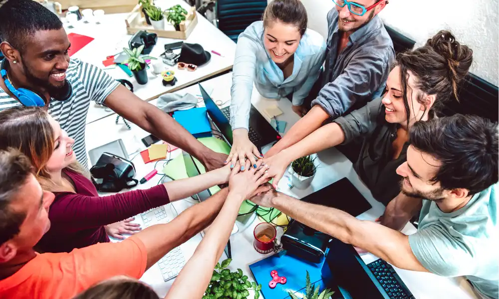 Grupo diverso de pessoas da geração Z colaborando em um ambiente de trabalho moderno, unindo as mãos em sinal de união e trabalho em equipe, rodeados por laptops e plantas.