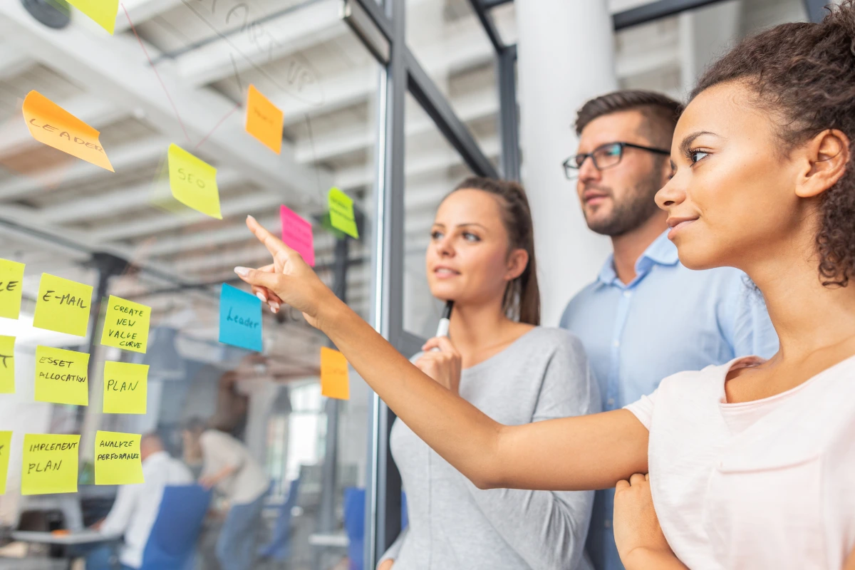 Grupo de três pessoas discutindo ideias durante uma reunião de trabalho, usando design thinking e post-its coloridos em um quadro de parede. Ambiente corporativo com foco em planejamento e estratégia.