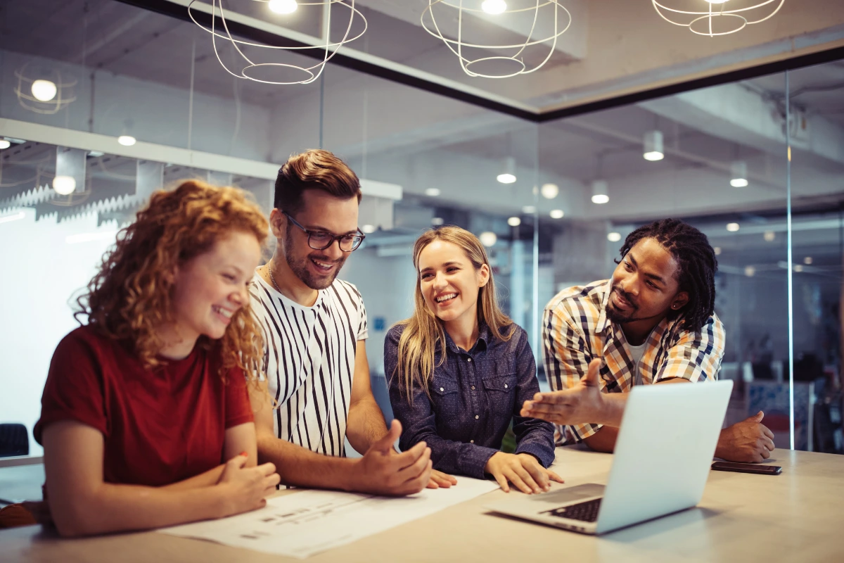 Grupo de jovens profissionais reunidos e sorrindo em torno de um notebook, em ambiente colaborativo que simboliza os pilares da liderança adaptativa.