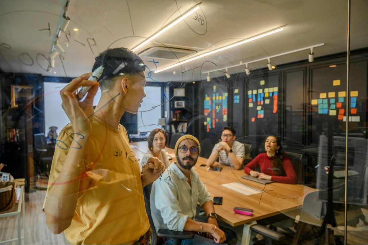 Reunião de equipe de negócios com uma mulher apresentando ideias para um grupo de colegas de trabalho em uma sala moderna e bem iluminada, com quadro de anotações e computador.