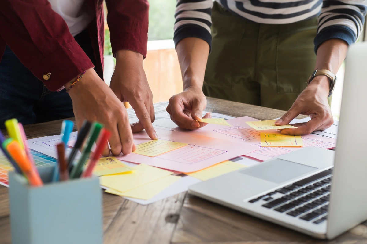 Duas pessoas trabalhando juntas em um projeto de design ou planejamento de treinamento com metodologia ativa, com vários papéis coloridos e um computador portátil na mesa.