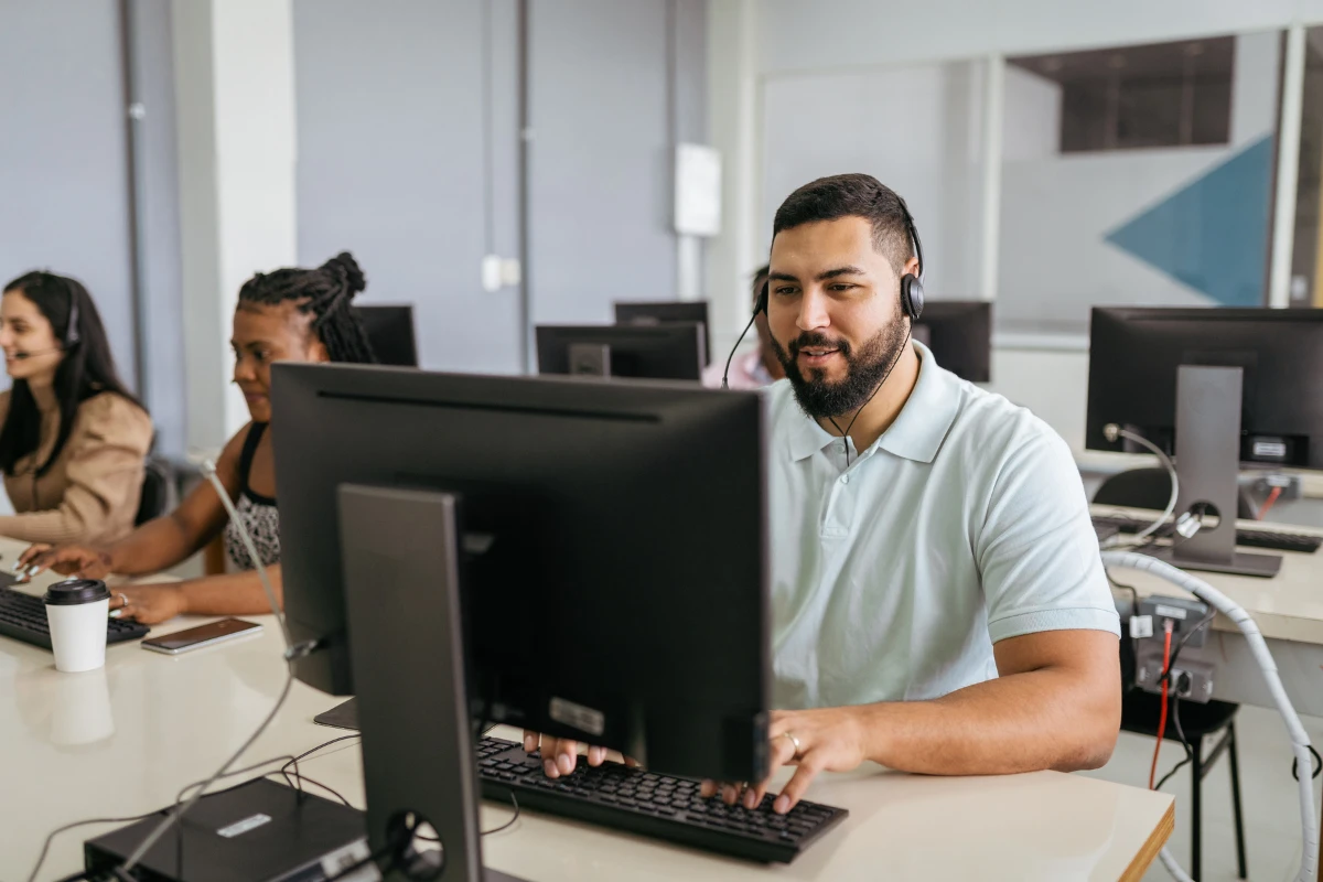 Homem com fone de ouvido sorrindo enquanto trabalha em um computador em ambiente de call center com colegas ao fundo. Usa computador como se estivesse organizando a área de customer education