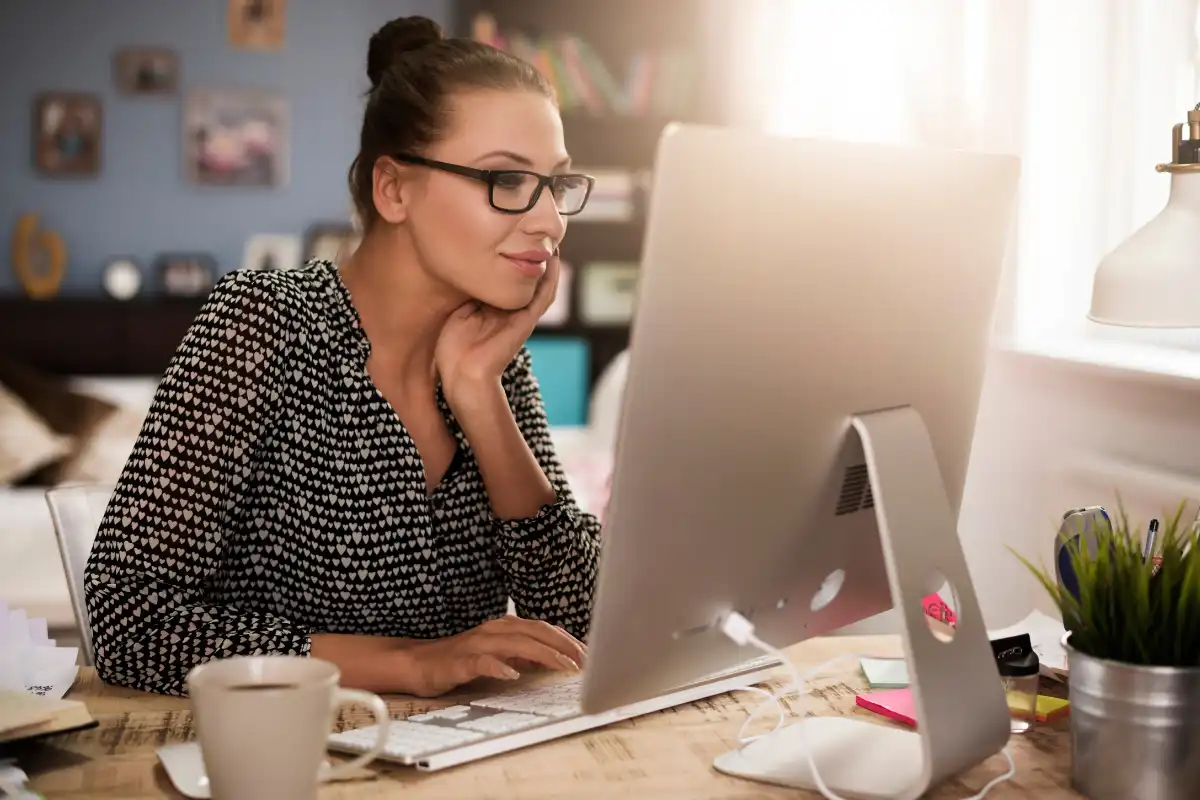 Mulher sorridente usando computador em um escritório doméstico, com foco e atenção na tela, ambiente organizado com livros e planta ao lado.