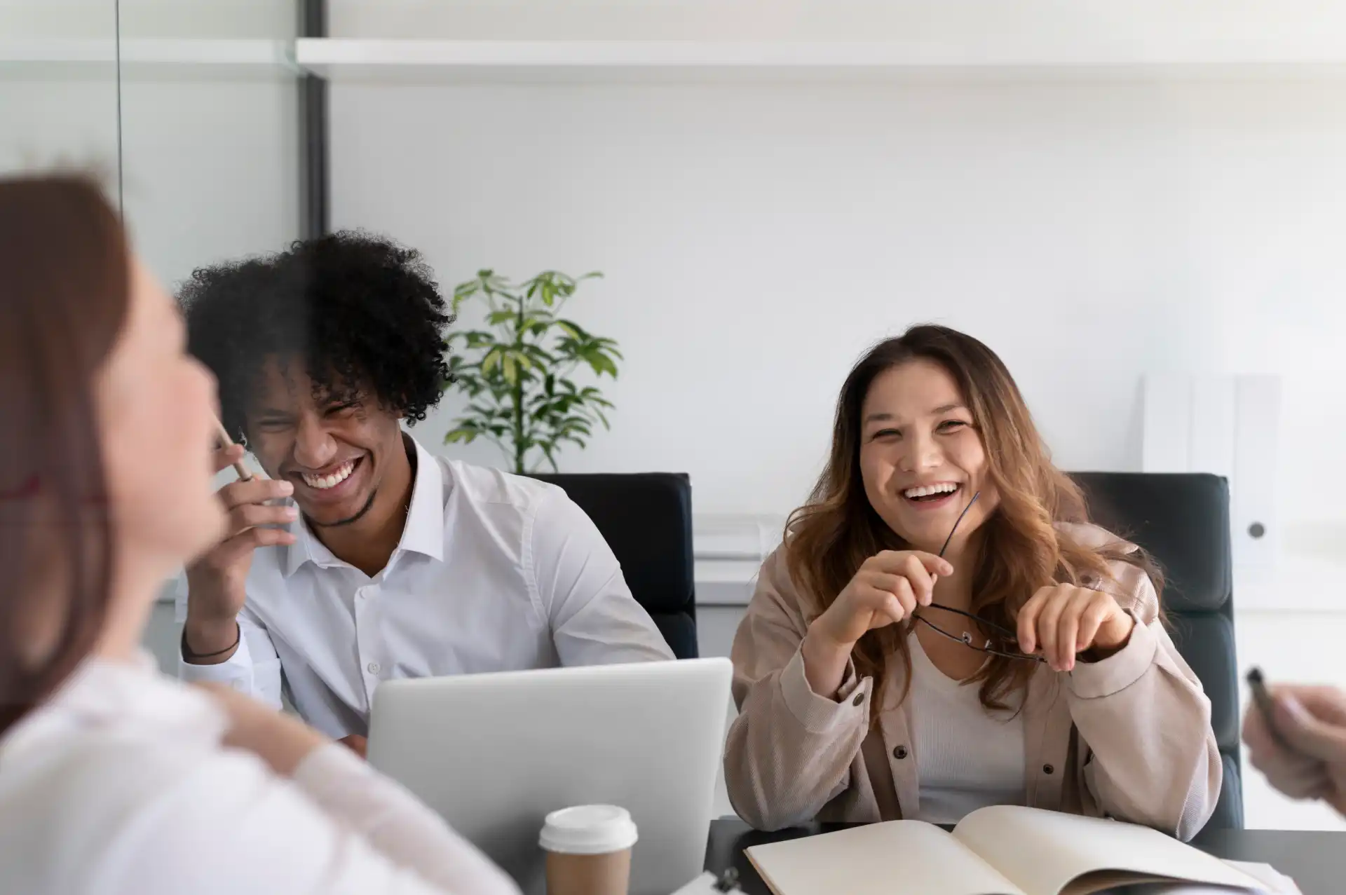 Grupo de colegas de trabalho sorrindo e conversando durante uma reunião em uma sala de escritório moderna, com um laptop, caderno e café na mesa.