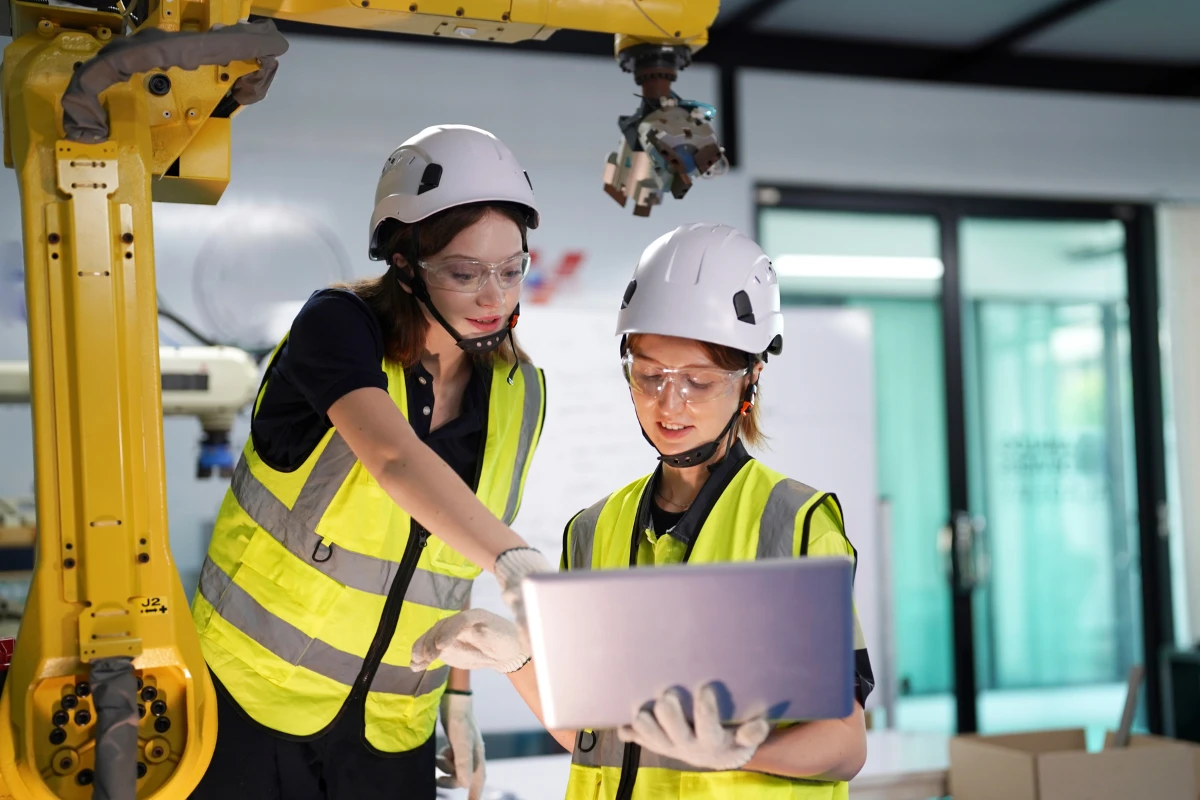 Engenheiras em uma fase de planejamento industrial usando capacetes de segurança e vestuário de alta visibilidade, ao lado de um robô na fábrica.