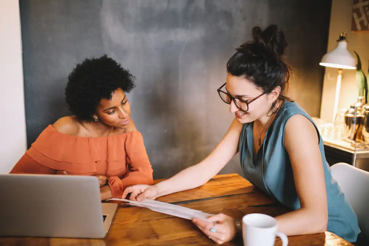 Duas mulheres sorridentes conversando e revisando um documento em uma mesa de escritório, com laptop, xícara e iluminação aconchegante ao fundo, representando colaboração e trabalho em equipe.