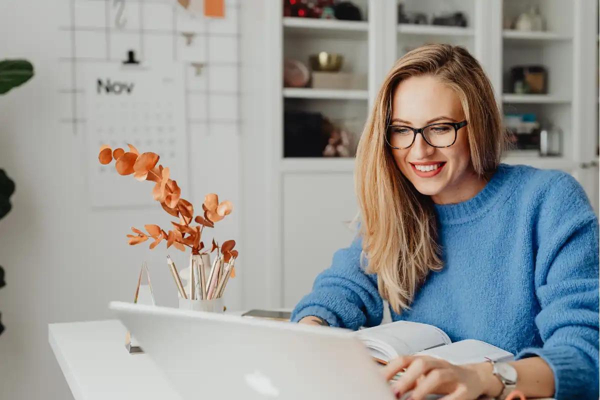 Jovem mulher de óculos e cabelo loiro claro trabalha em casa usando laptop e aproveita um momento de estudo com sorriso e ambiente organizado ao fundo.