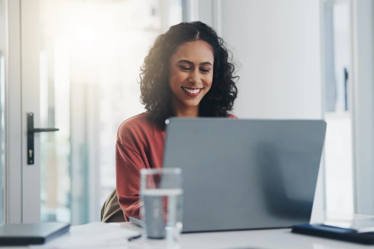 Mulher sorridente trabalha em um escritório e usa um notebook sobre a mesa, com um copo ao lado, em ambiente claro e bem iluminado
