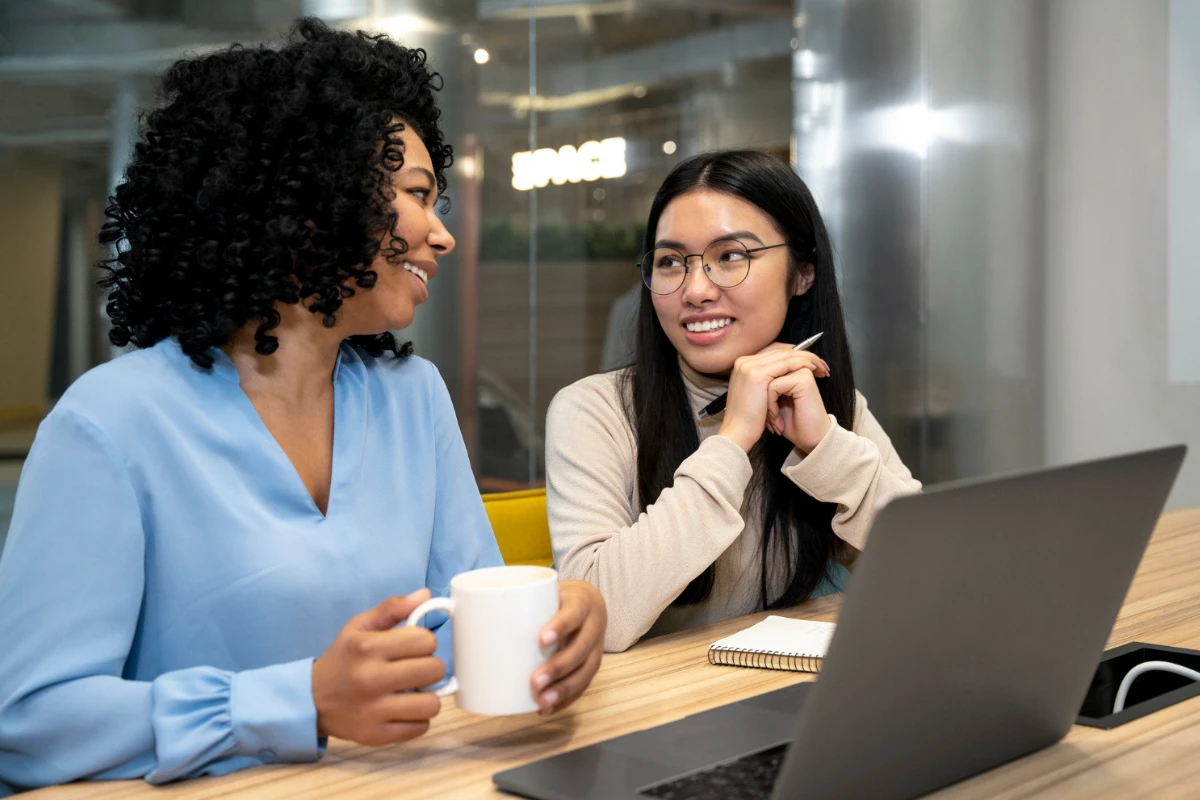 Duas mulheres em ambiente corporativo conversam em uma mesa de trabalho, com uma delas segurando uma caneca e a outra sorrindo diante de um notebook, em escritório com luz natural.