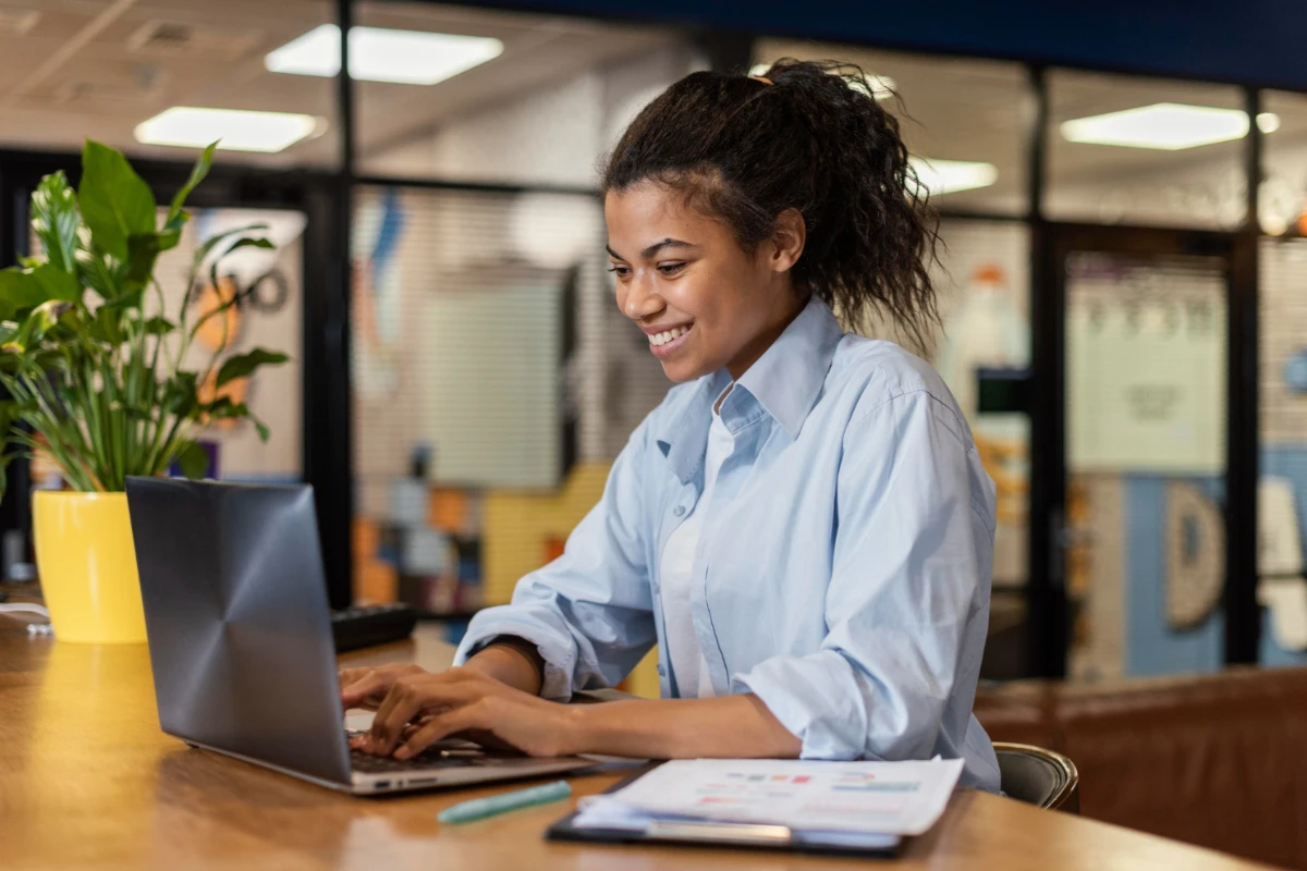 Jovem profissional sorrindo enquanto digita no notebook em um escritório moderno, com planta em vaso e documentos sobre a mesa. Ambiente de trabalho colaborativo.