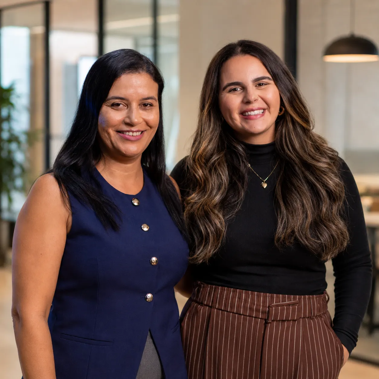 Duas mulheres sorrindo em foto corporativa, uma com vestido azul sem mangas e a outra com blusa preta de manga longa e saia listrada, em ambiente de escritório com fundo desfocado.
