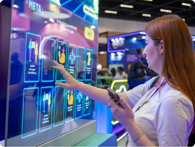Mulher aponta para tela em exposição de tecnologia, com gráficos e blocos luminosos de aplicativo ou sistema em um estande de feira, em ambiente interno.