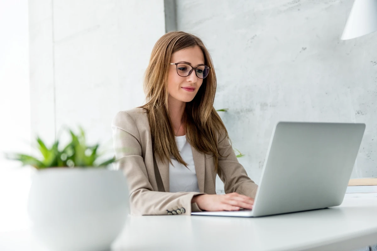 Mulher profissional com óculos sentada em escritório moderno, usando notebook em uma mesa branca, com parede de concreto e luz natural ao fundo.
