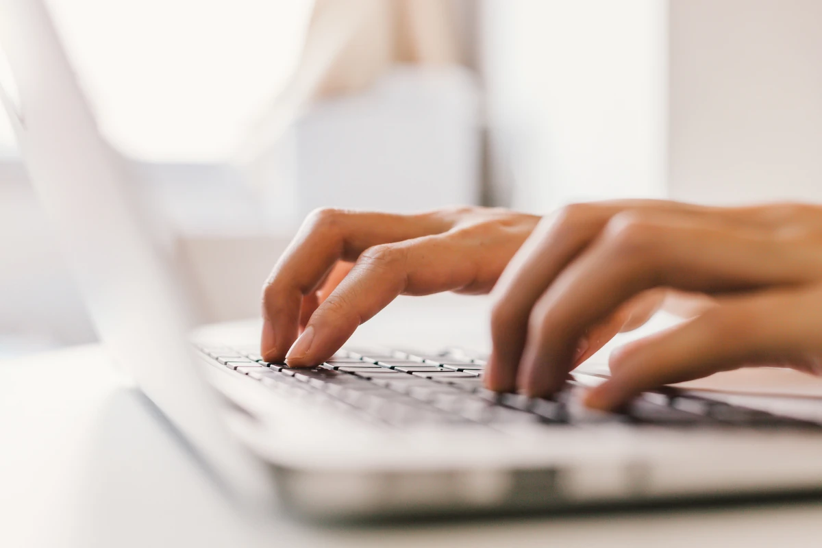Pessoa digitando em um teclado de computador em ambiente de escritório, com as mãos visíveis e foco na ação de escrever, cenário claro e desfocado ao fundo.