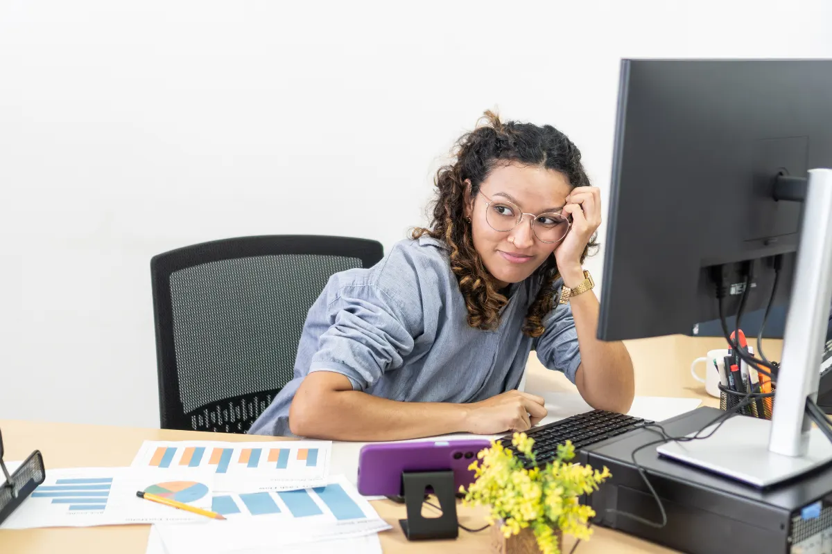 Mulher com óculos sentada em uma mesa de escritório usando computador e analisando gráficos em uma reunião, com celular e pastas ao lado.