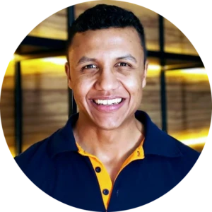 Retrato sorridente de um homem jovem com camisa azul e detalhe amarelo, em ambiente interno com iluminação quente ao fundo.